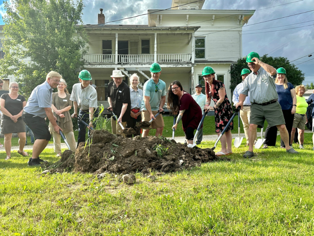 A group of people wearing green helmets break ground with shovels in front of a white house, while others observe in the background.